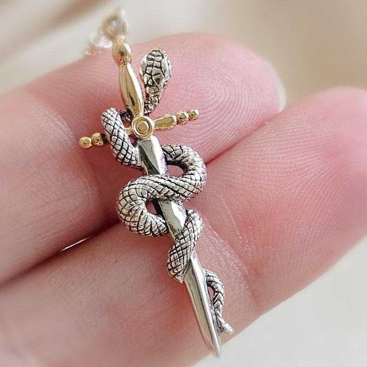 Close-up of the Snake & Dagger Necklace resting on soft ivory fabric beside a glass bowl. The pendant features a silver serpent coiled around a gold dagger, highlighting the mix of textures and metals in an elegant, minimalist setting.