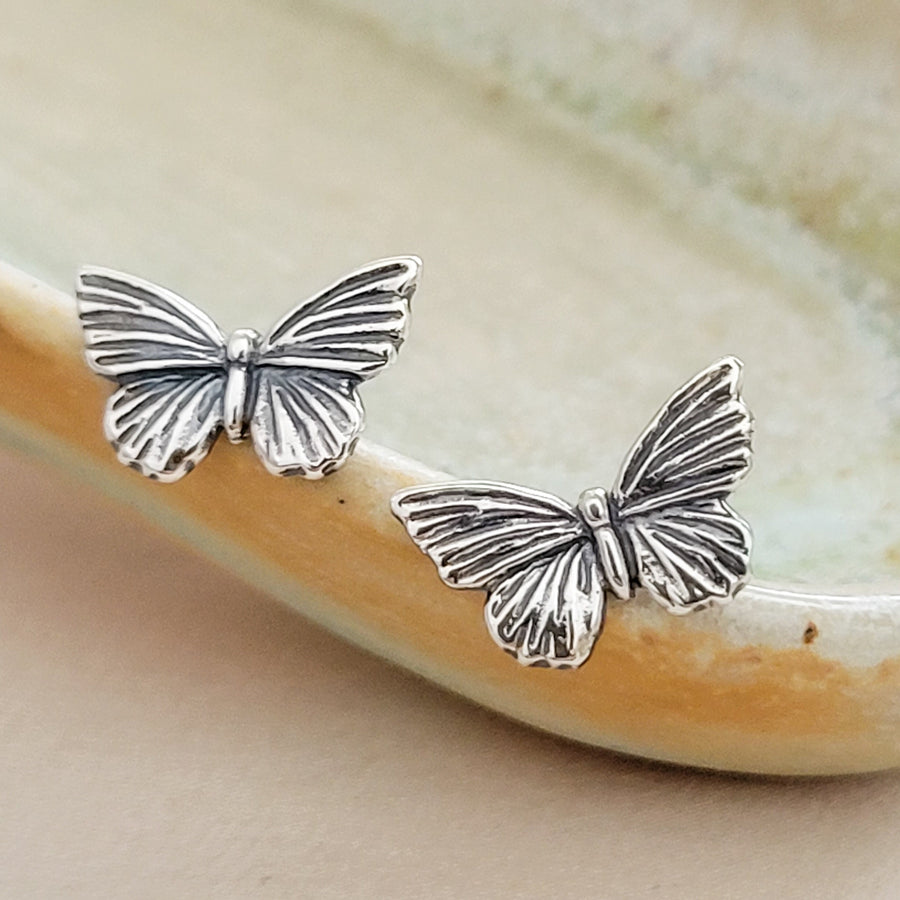 Pair of Sterling silver Butterfly studs earrings shown on the rim of a sage green and tan ceramic dish, by Studio Blue on Etsy