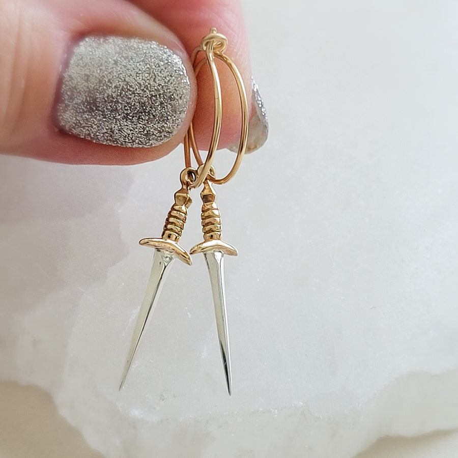 Bronze & Sterling Silver Dagger charm on a 15mm Gold fill hoop earring, shown being held between 2 fingers on a white background. By Studio Blue on Etsy